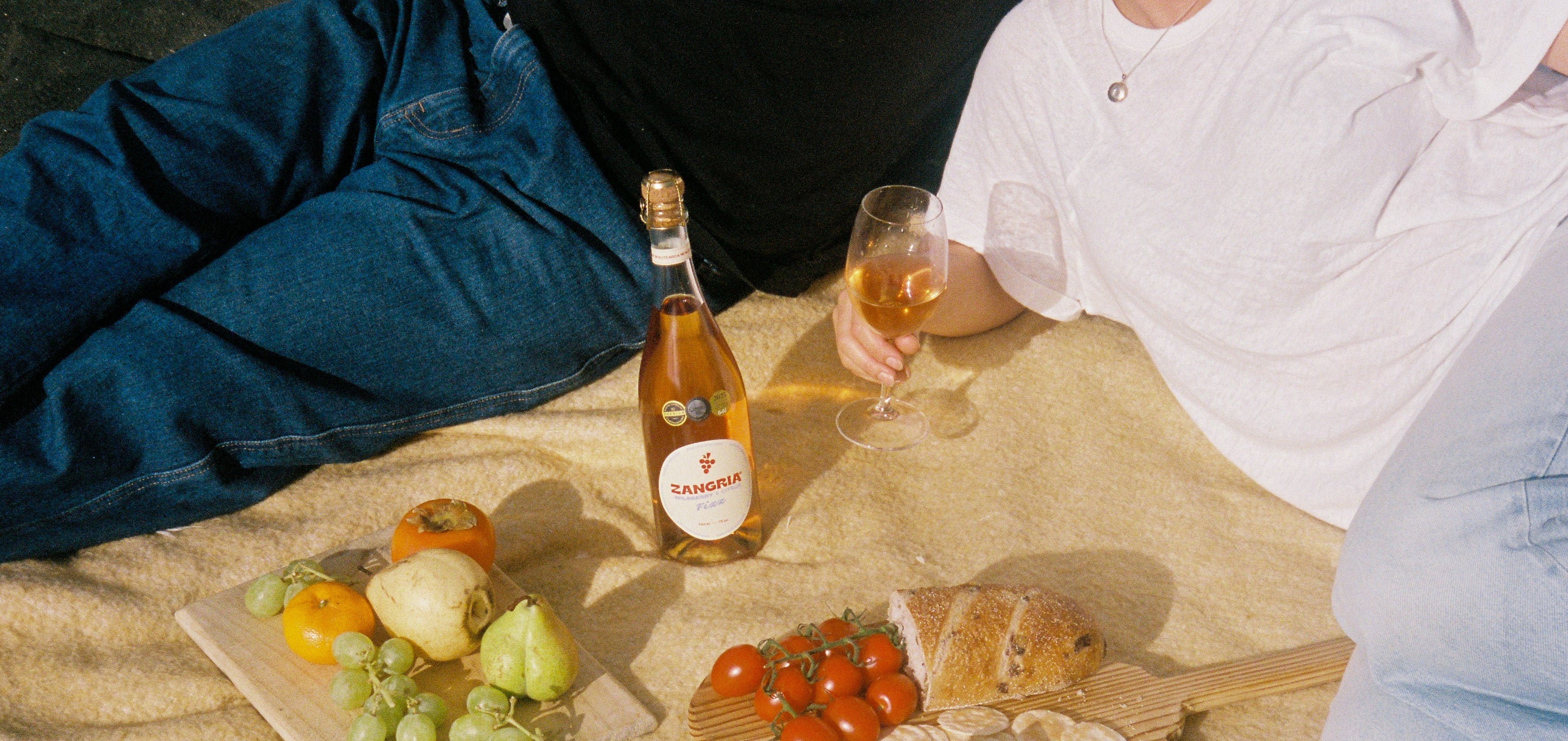 A lifestyle picnic scene on a beige blanket featuring a bottle of Valtaris sparkling wine, a glass of bubbly pinkish-amber liquid, fresh fruit, sliced bread, and tomatoes, with two people partially visible in blue jeans and a white t-shirt.