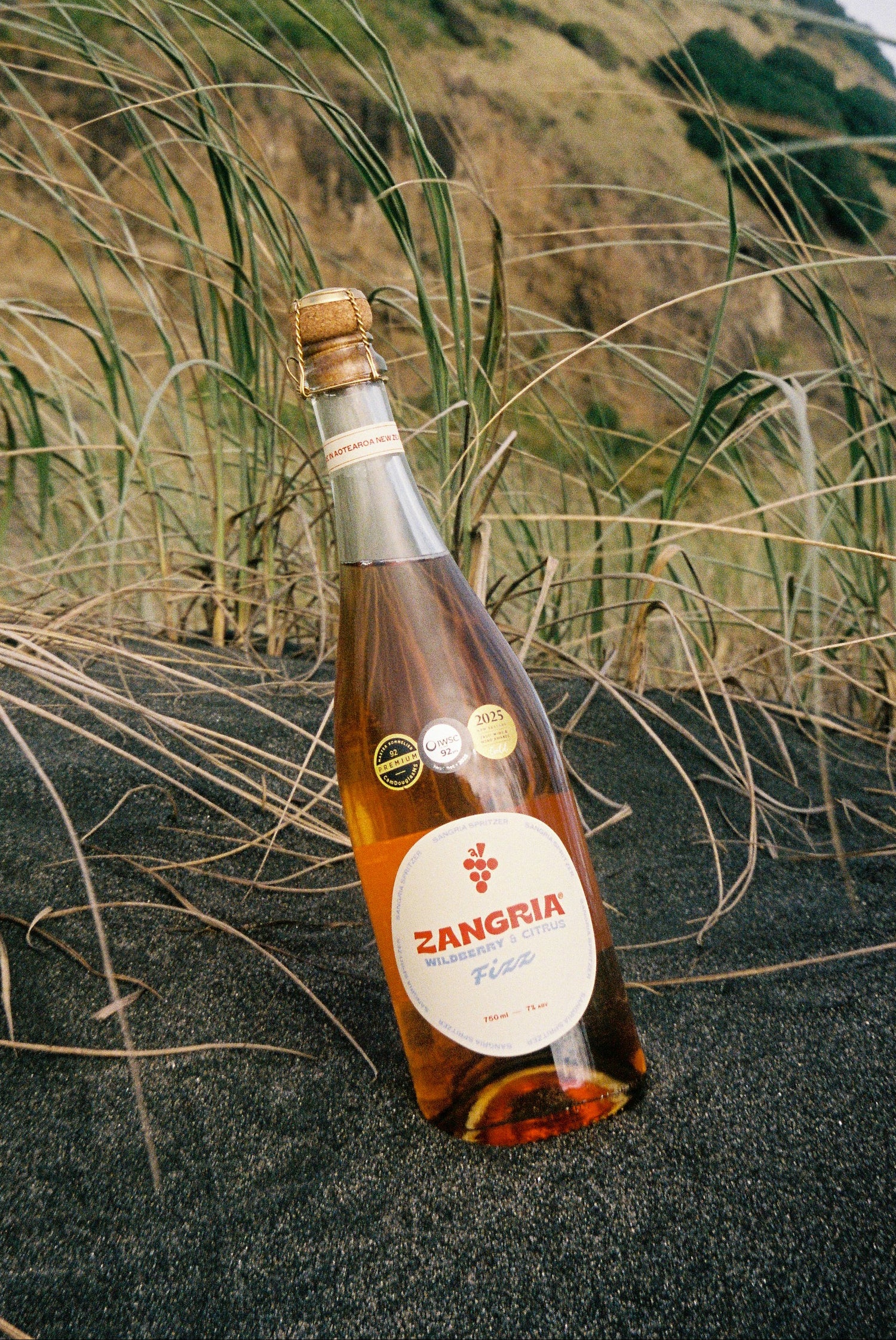 A bottle of Zangria Fuego tequila on the rocks with a cork stopper, set on dark sand amidst tall beach grass with a grassy hill in the background.