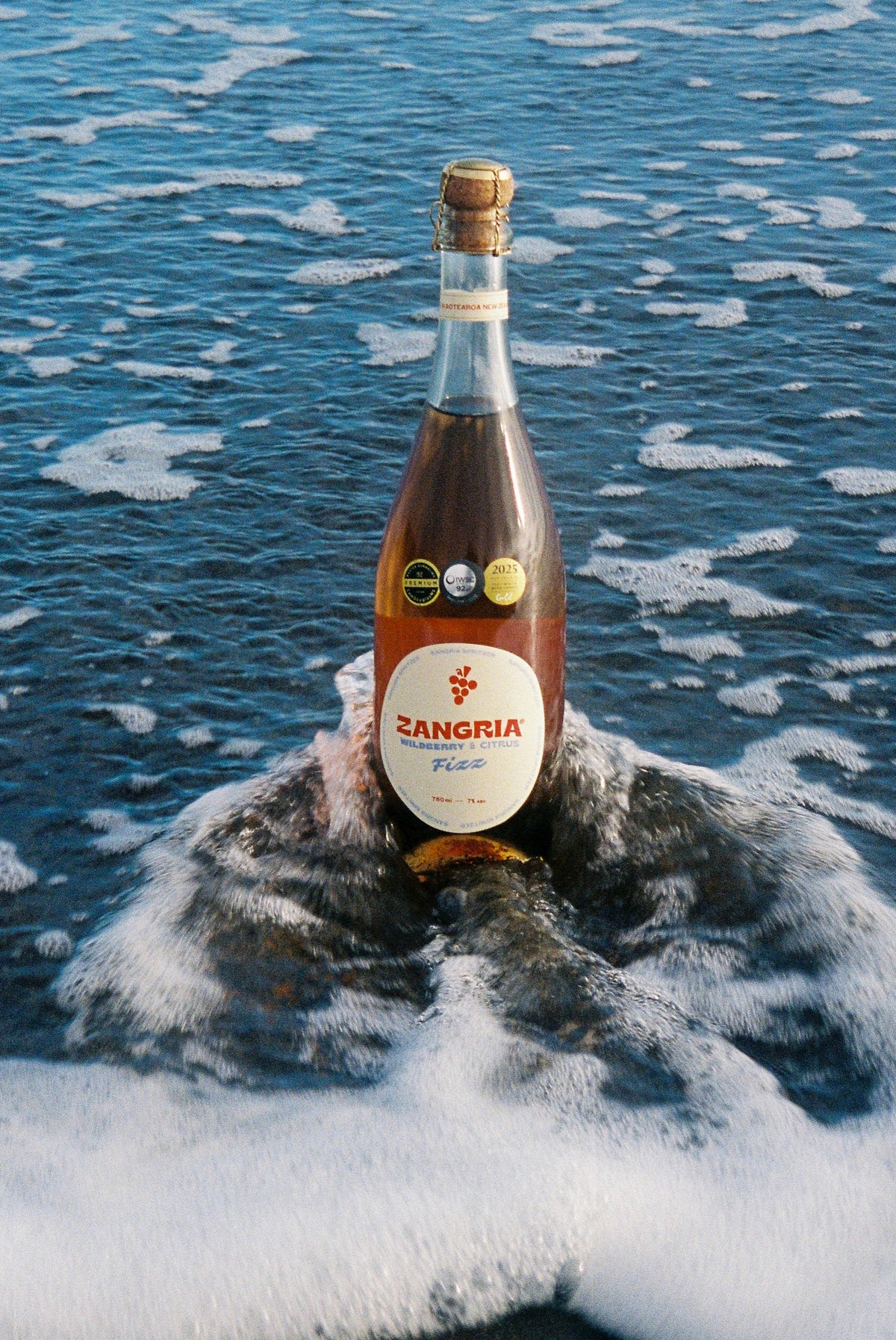 A bottle of Zangria Flor sparkling wine stands upright in foamy ocean water, with blue sea and scattered ice visible around it.